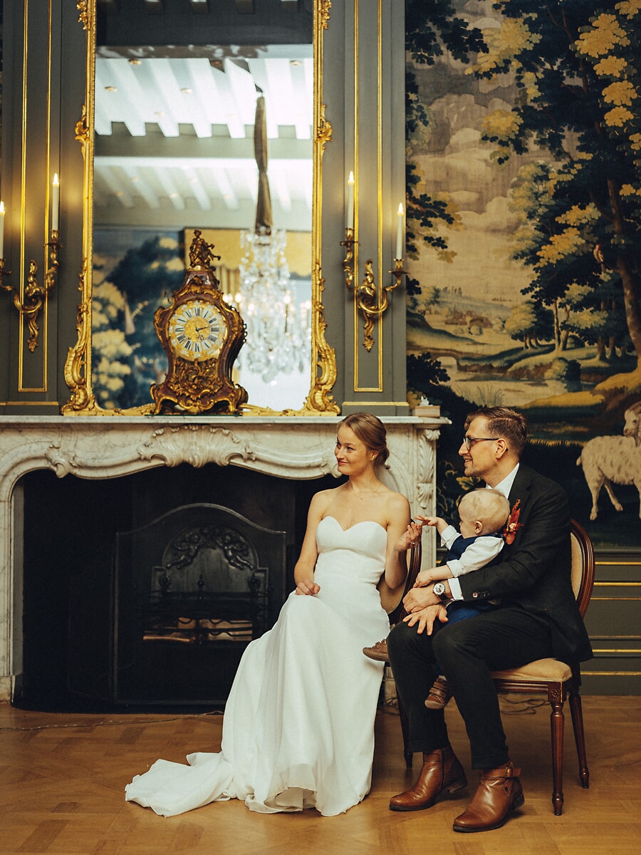 Wedding Ceremony SP24 Bruid, bruidegom en hun baby zitten voor een sierlijke vergulde spiegel en schouwklok, een elegant bruidsportret van het burgerlijk huwelijk