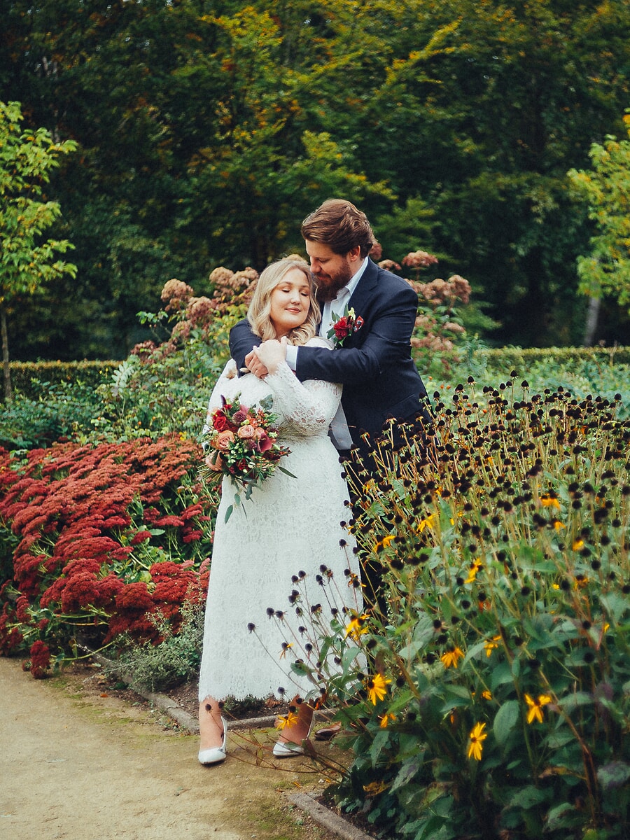 Wedding Ceremony SP27 Bruidegom omhelst zijn bruid van achteren in een herfsttuin vol rode bloemen, haar boeket in de hand, door een trouwfotograaf in Amsterdam