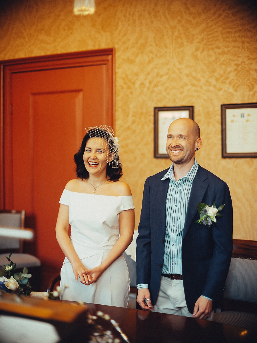 Wedding Ceremony SP28 Lachende bruid in een schoudervrije jurk en korte sluier naast haar bruidegom in een Amsterdams stadhuis, vastgelegd door een trouwfotograaf in Amsterdam