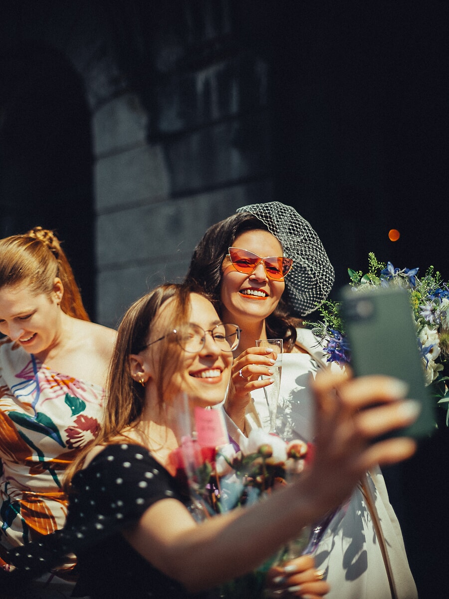 Wedding Ceremony SP29 In een korte sluier en cat-eye zonnebril maakt de bruid een selfie met lachende gasten, een spontane trouwfoto