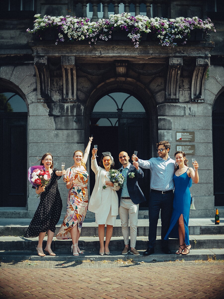 Wedding Ceremony SP30 Op de trap van een met bloemen versierd gemeentehuis proosten het bruidspaar en de gasten, vastgelegd door een trouwfotograaf in Amsterdam