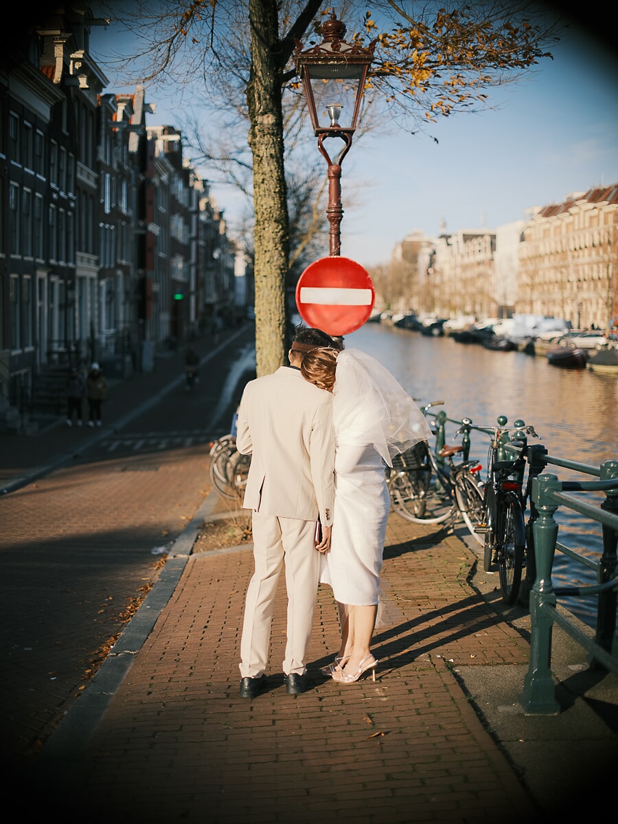 Wedding Ceremony SP4 Stel in crèmekleurige trouwkleding loopt hand in hand door een zonovergoten Amsterdamse grachtenstraat langs geparkeerde fietsen, vastgelegd door een trouwfotograaf in Amsterdam