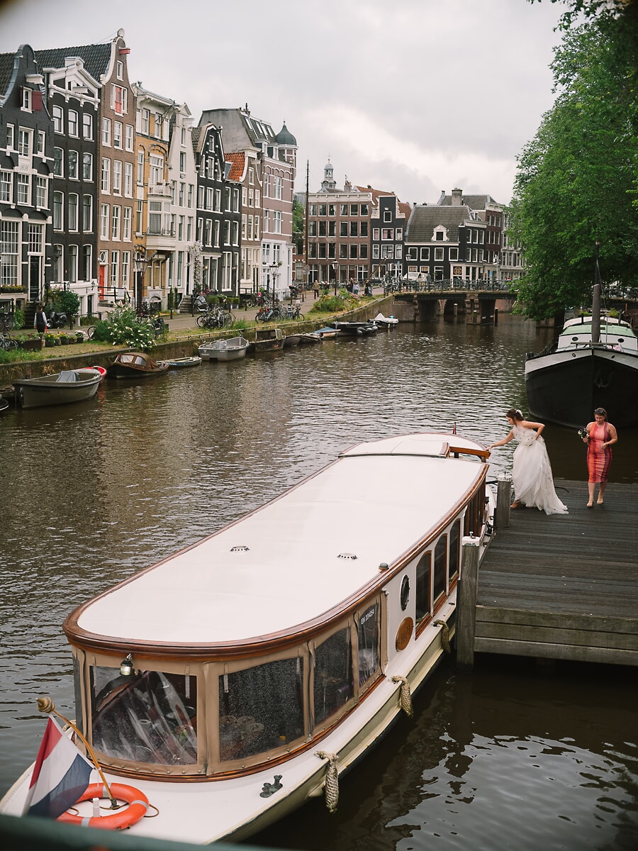 Wedding Ceremony SP7 Witte salonboot afgemeerd langs een gracht met historische panden terwijl de bruid aan boord stapt, een weids beeld uit de Amsterdamse trouwfotografie