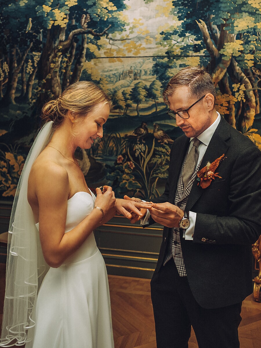 Wedding Ceremony SP34 Bruid schuift de ring om de vinger van haar bruidegom voor een groot boswandtapijt in een zaal met visgraatparket, een teder ceremoniemoment