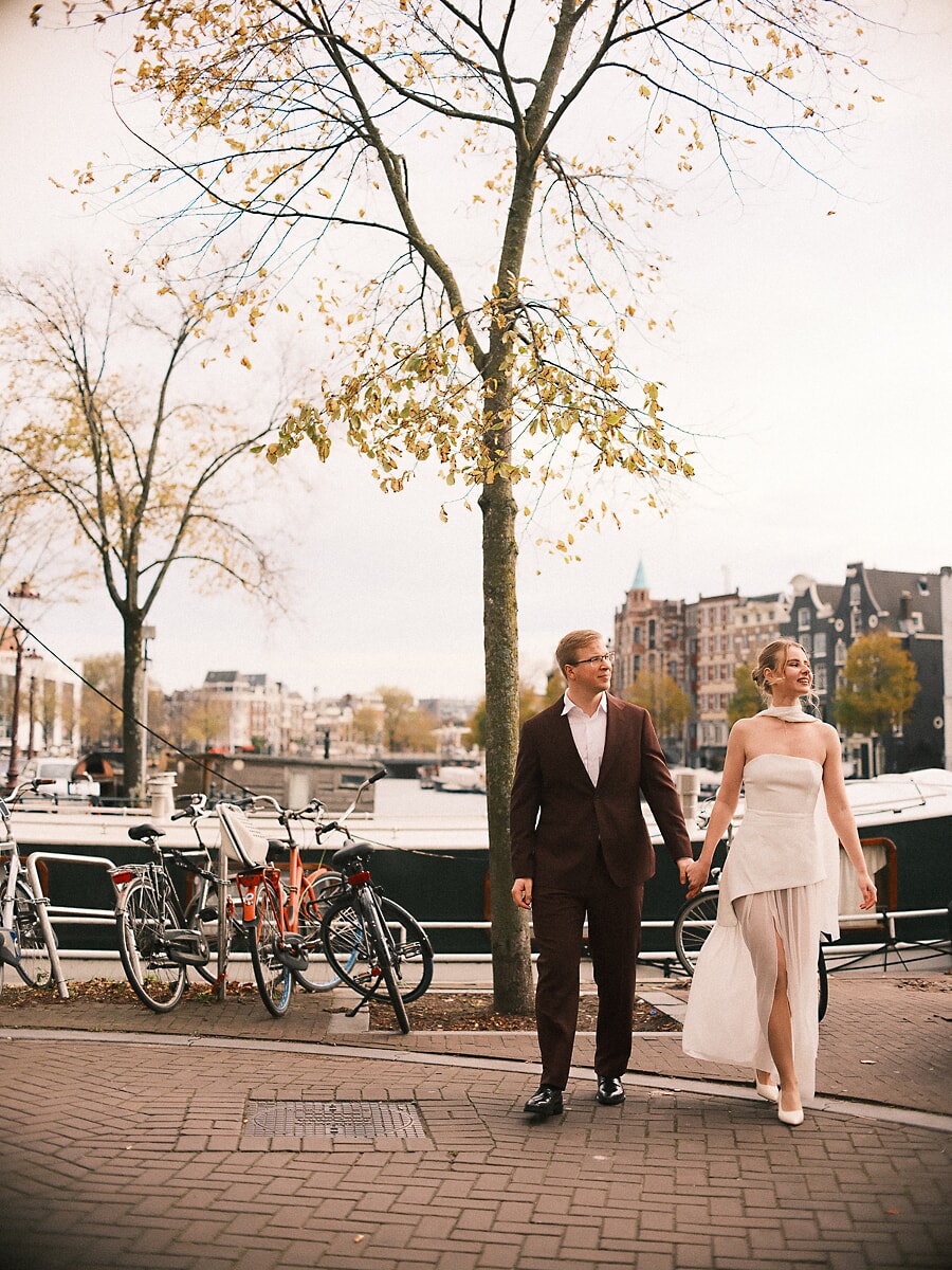 Wedding Bridal SP10 Bruidegom in bruin ribfluweel en bruid in geplooide ivoren rok lopen onder een herfstboom langs de Amstel, een ontspannen foto