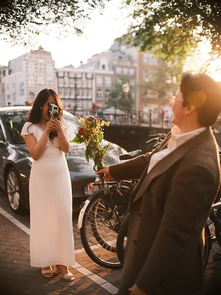 Wedding Bridal SP11 Bruid filmt haar lachende bruidegom met een oude filmcamera langs een gracht, een shoot met een trouwfotograaf in Amsterdam