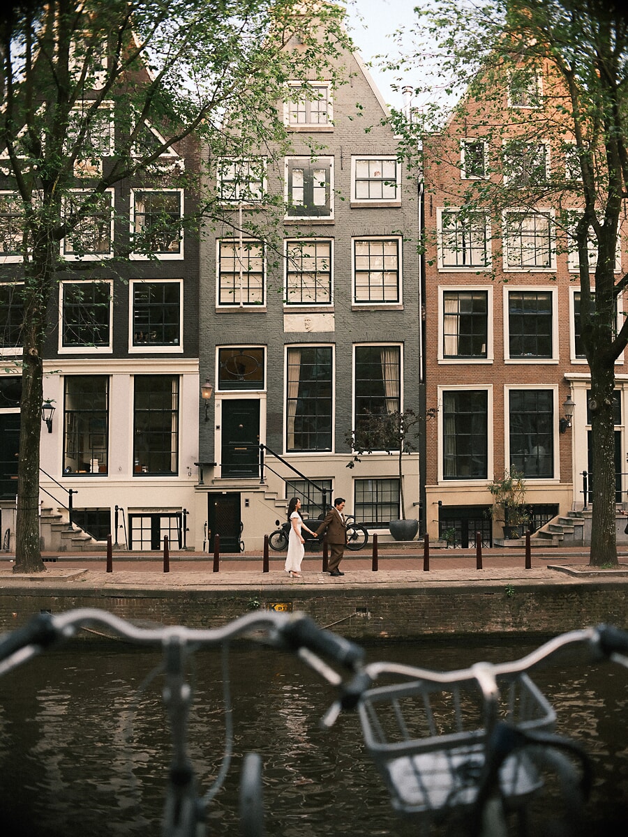 Wedding Bridal SP16 Langs onscherpe fietssturen wandelen de pasgetrouwden over de kade onder hoge grachtenpanden, een documentaire trouwfoto