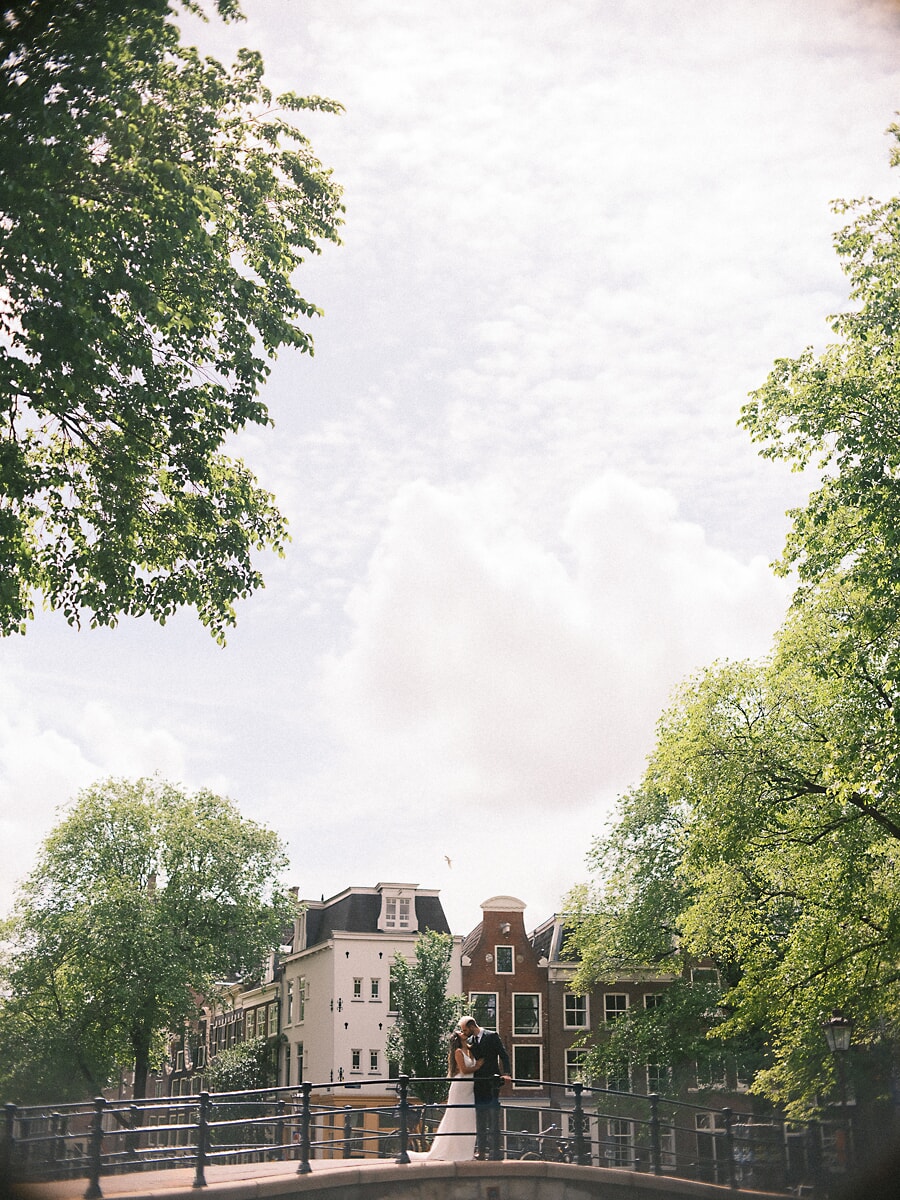Wedding Bridal SP2 Pasgetrouwden omhelzen elkaar op een witte grachtenbrug tussen hoge zomerbomen, een weids beeld uit hun bruidsshoot