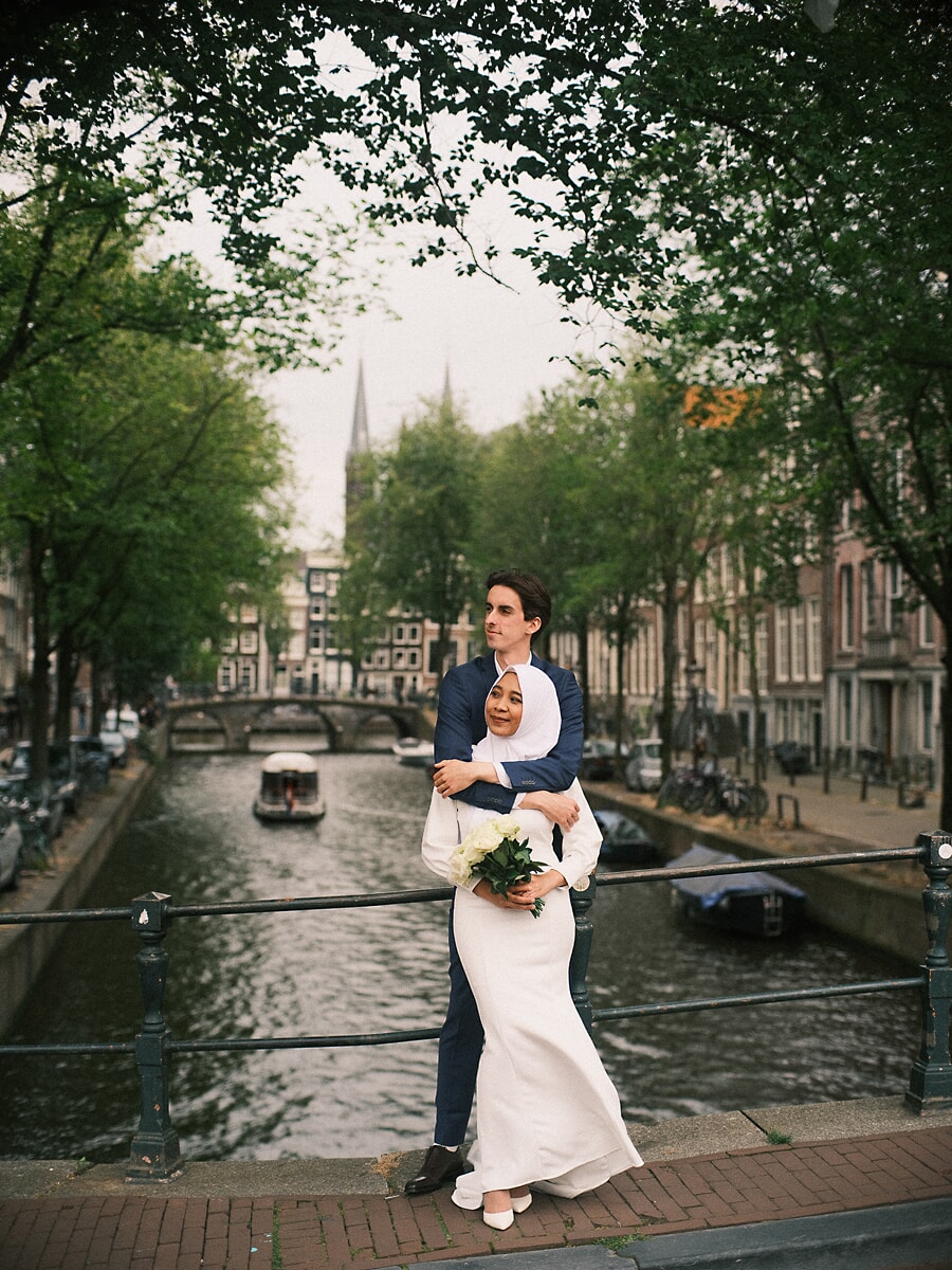 Wedding Bridal SP5 In ivoren jurk en hijab leunt de bruid in de armen van haar bruidegom op een brug, vastgelegd door een trouwfotograaf in Amsterdam