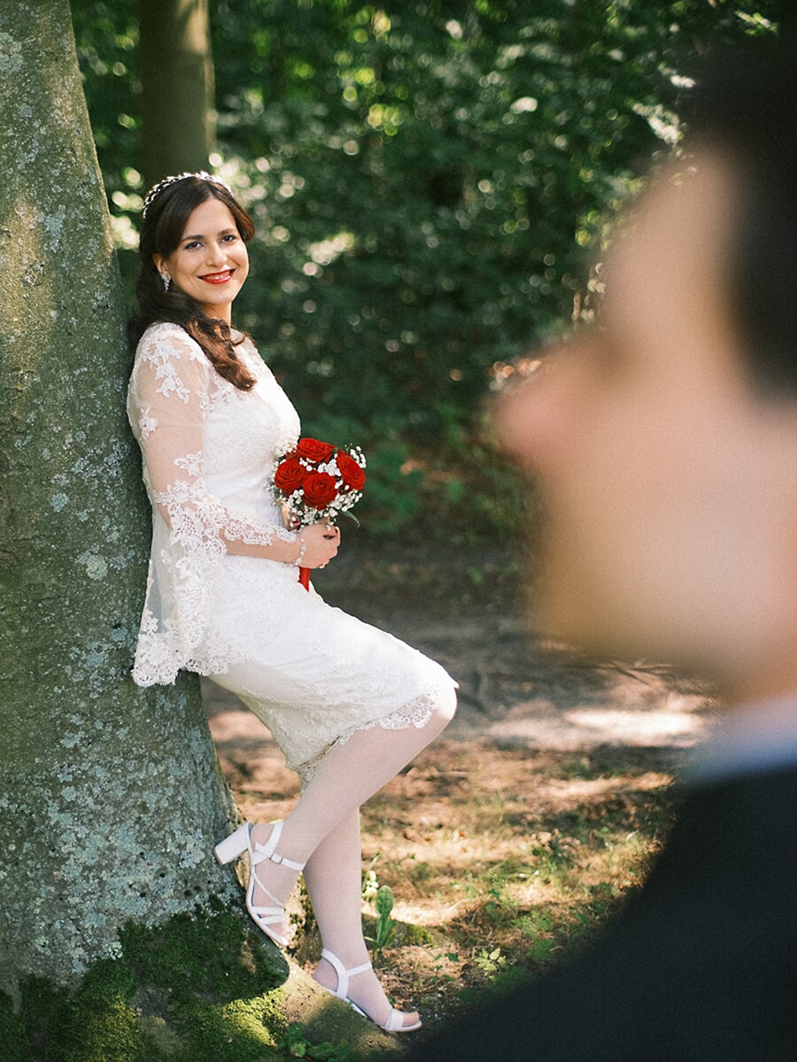 Wedding Bridal SP6 Bruid in kanten jurk leunt tegen een bemoste boom met rode rozen, haar bruidegom onscherp op de voorgrond, een romantische trouwfoto