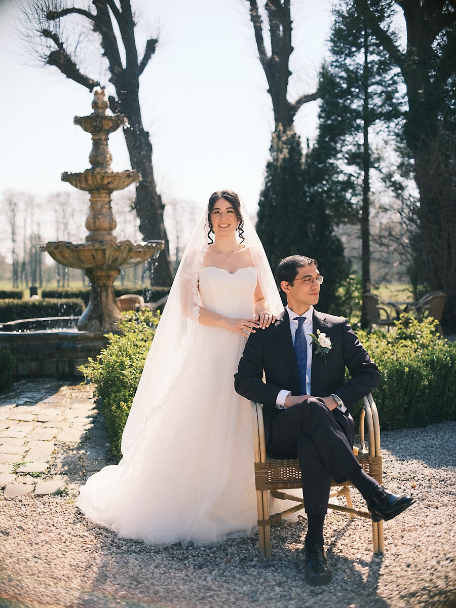 Wedding Bridal SP23 Bij een stenen fontein in een kale wintertuin legt de bruid haar hand op de schouder van haar zittende bruidegom, een elegant beeld