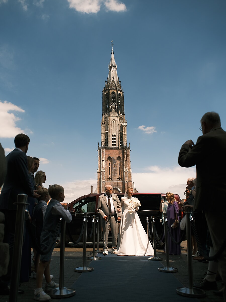 Wedding SP1 Een trouwfotograaf in Amsterdam legt vast hoe de bruid uit een donkere auto stapt op de blauwe loper voor de Nieuwe Kerk