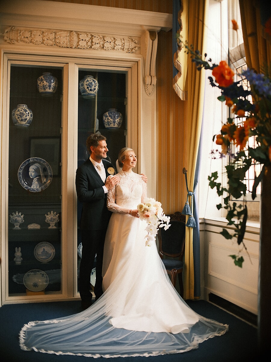 Wedding SP10 Pasgetrouwden poseren bij een kast vol Delfts blauw, de sluier uitgespreid over de vloer, een elegant trouwportret