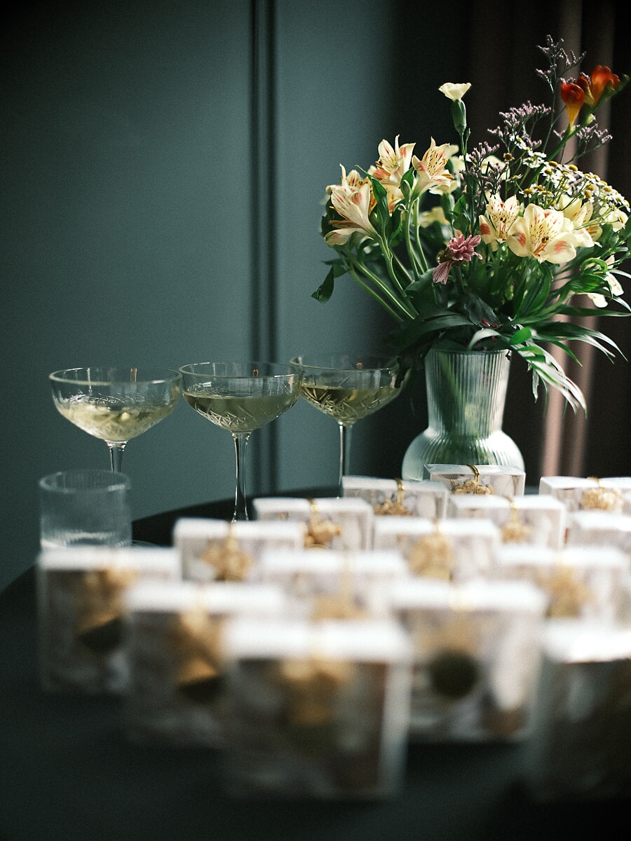 Wedding SP12 Champagnecoupes en glazen bedankjes rond een geribbelde vaas met bloemen op een donkere feesttafel, een gestileerd bruiloftsdetail