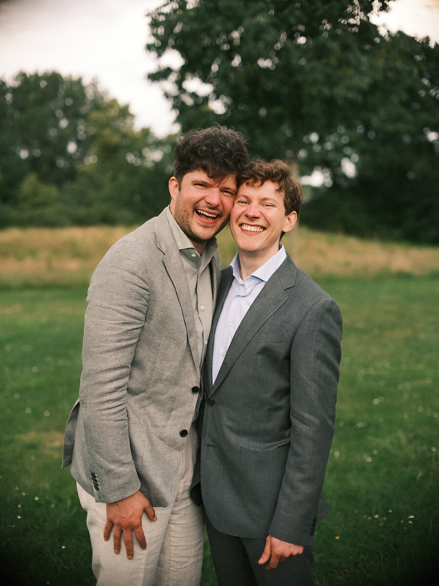 Wedding SP15 Twee bruidegoms in grijs en linnen lachen wang aan wang in een weiland, een LHBTI+ stel vastgelegd door een trouwfotograaf in Amsterdam