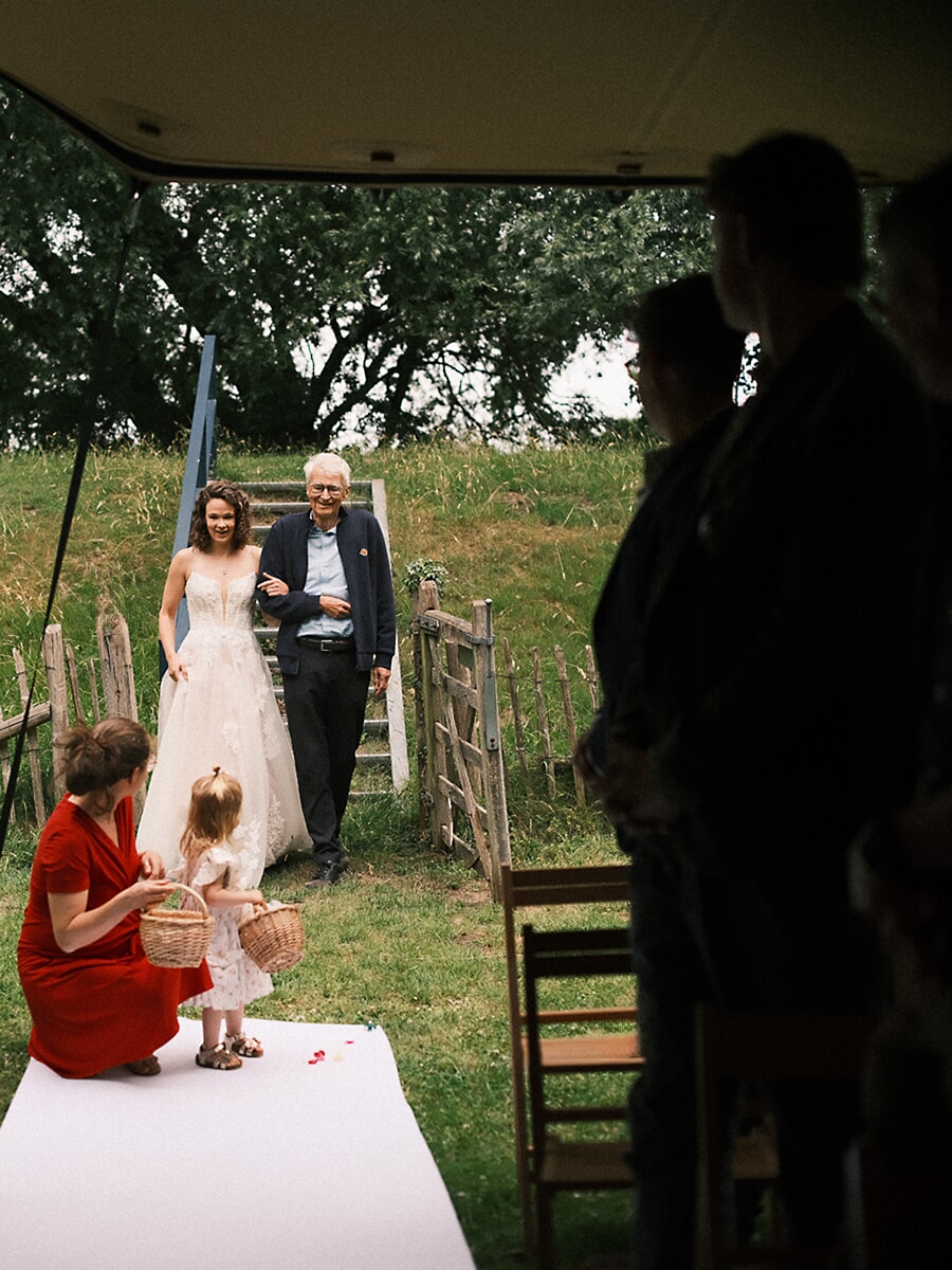 Wedding SP16 Aan de arm van haar vader loopt de bruid over de witte loper langs een bloemenmeisje dat blaadjes verzamelt, een spontane ceremoniefoto