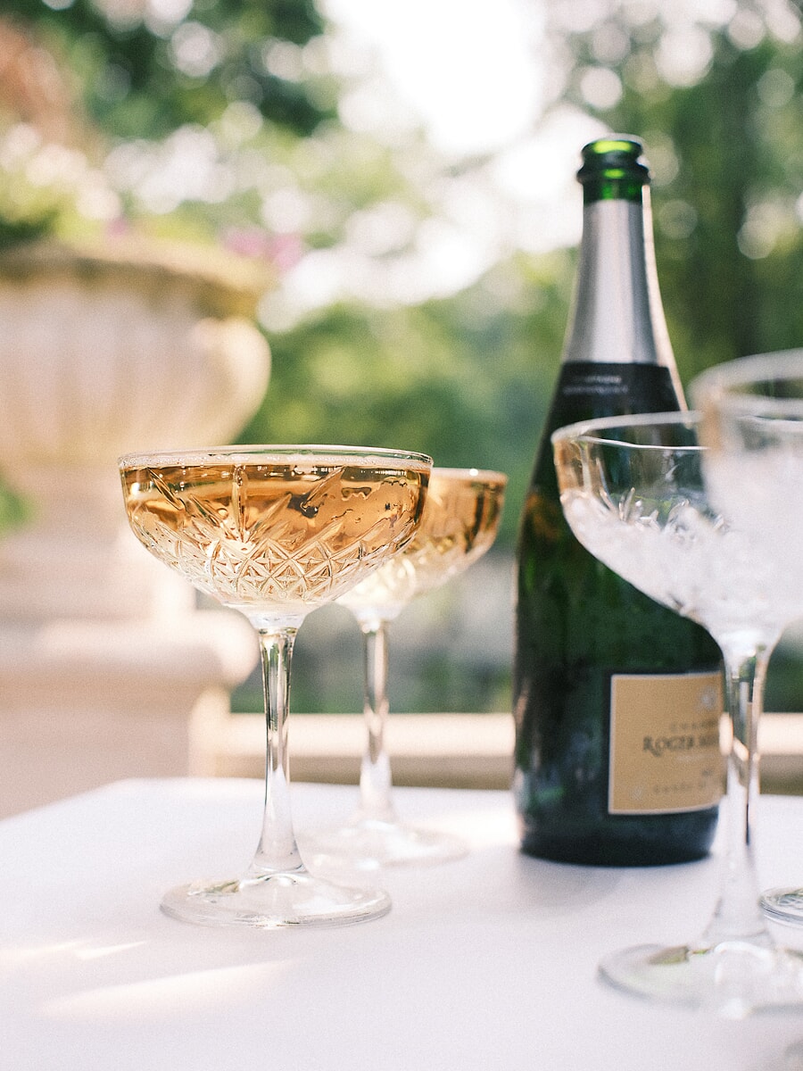Wedding SP17 Gekoelde fles champagne naast twee gevulde kristallen coupes op een zonnig terras, een feestelijk detail uit de bruiloftsfotosessie