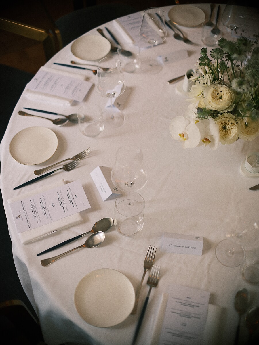 Wedding SP18 Bovenaanzicht van een ronde tafel met wit linnen, menukaarten, zilveren bestek en lichte ranonkels, een verfijnd bruiloftsdetail