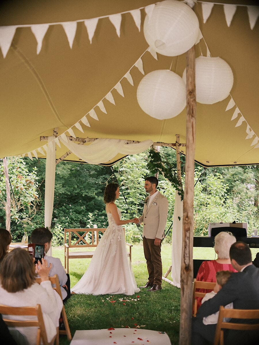 Wedding SP19 Onder lampionnen en vlaggetjes in een tuintent staan ze hand in hand voor hun gasten, vastgelegd door een trouwfotograaf in Amsterdam