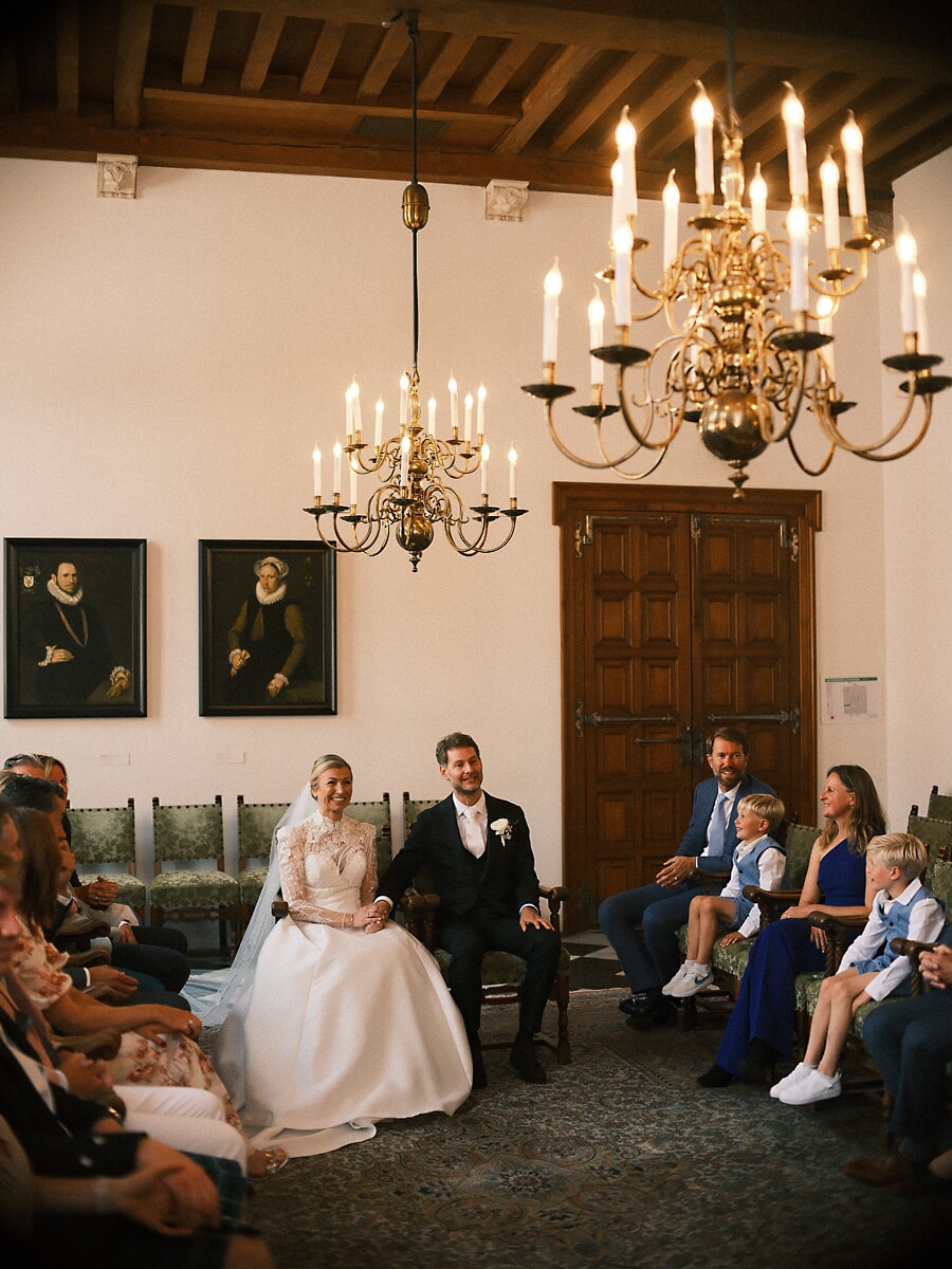 Wedding SP2 Pasgetrouwden zitten naast elkaar onder koperen kaarsenkronen terwijl familie en kinderen toekijken, een natuurlijke huwelijksfoto