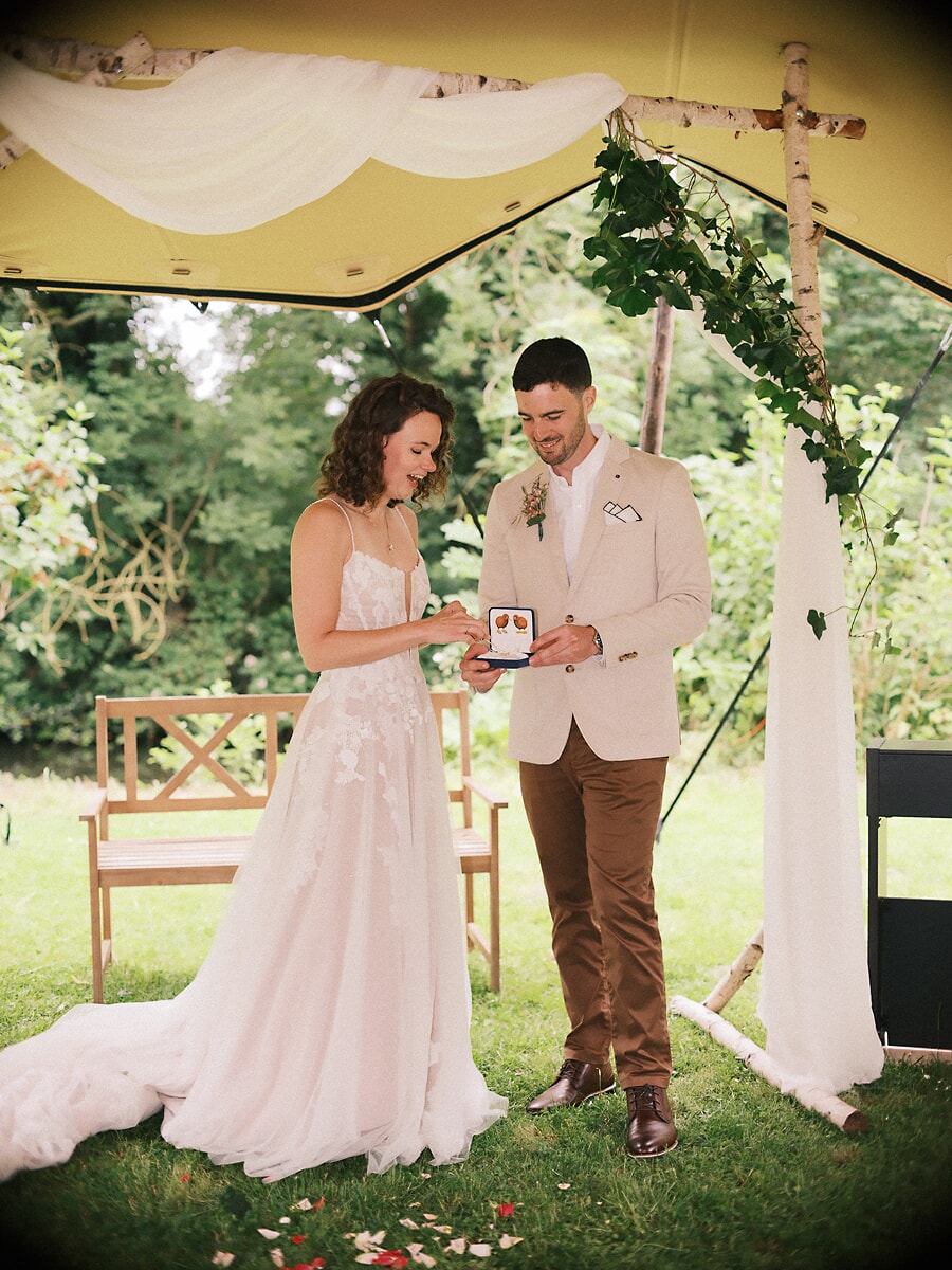 Wedding SP21 Bij het wisselen van de ringen onder een berkenboog lachen ze naar het geopende ringdoosje, vastgelegd door een trouwfotograaf in Amsterdam