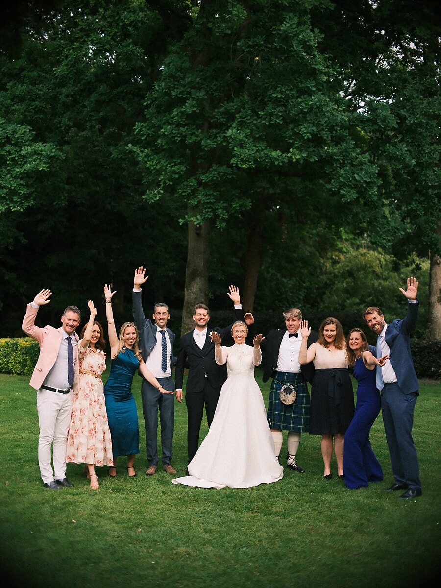 Wedding SP22 Het bruiloftsgezelschap gooit de handen in de lucht op het gazon, een gast in kilt naast de stralende bruid, een levendige groepsfoto