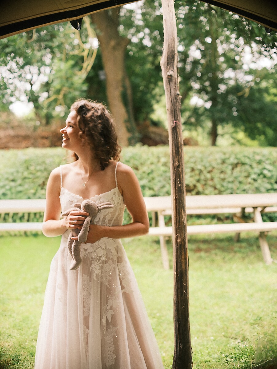 Wedding SP23 Lachend onder de rand van de tent houdt de bruid in een jurk met spaghettibandjes een knuffelkonijn vast, een vrolijk trouwportret