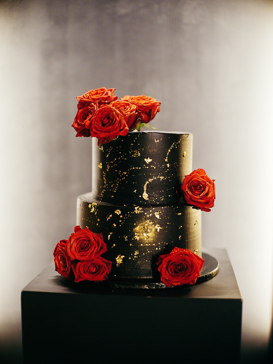 Wedding SP24 Zwart met gouden taart van twee lagen met rode rozen op een donkere sokkel, een opvallend detail uit de bruiloftsfotografie
