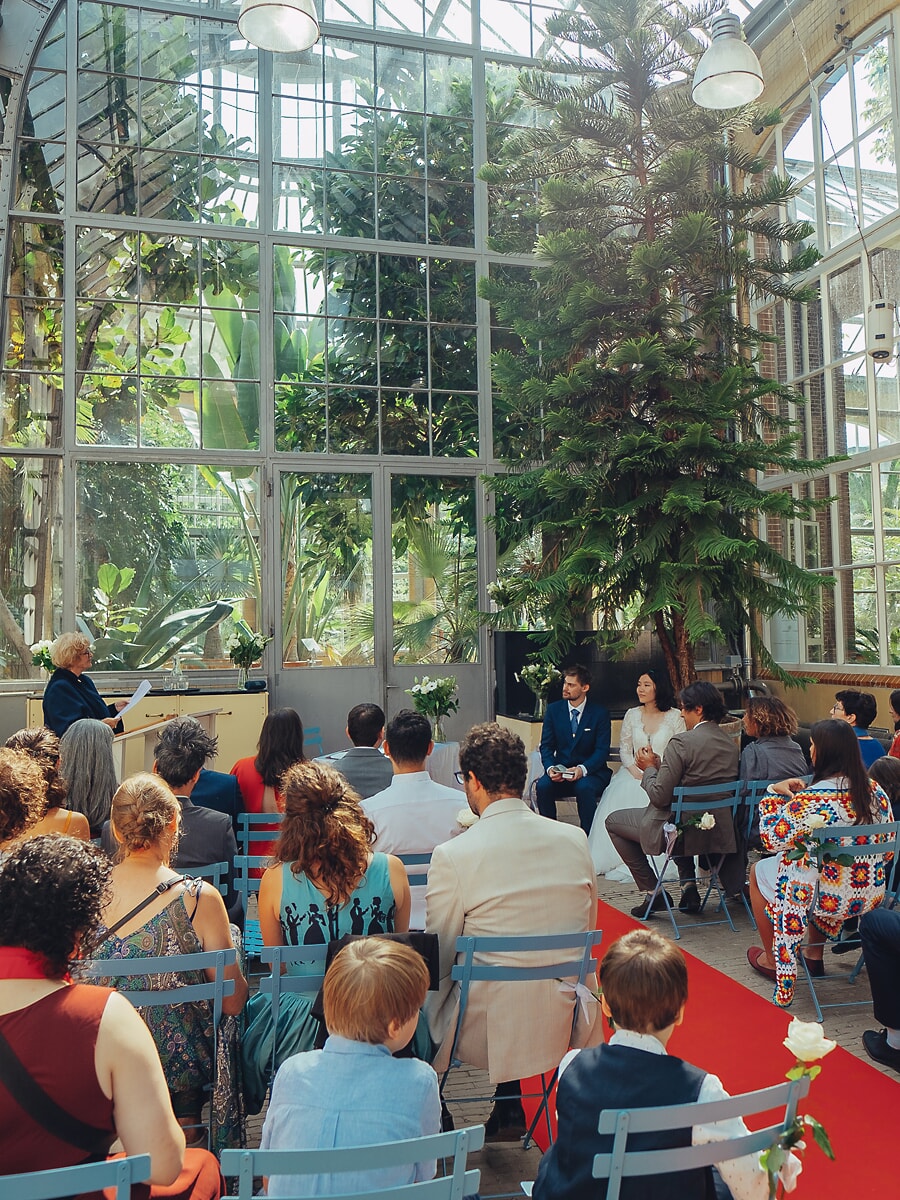 Wedding SP25 Gasten op blauwe stoelen volgen de ceremonie in een glazen orangerie, met rode loper naar het stel, van een trouwfotograaf in Amsterdam