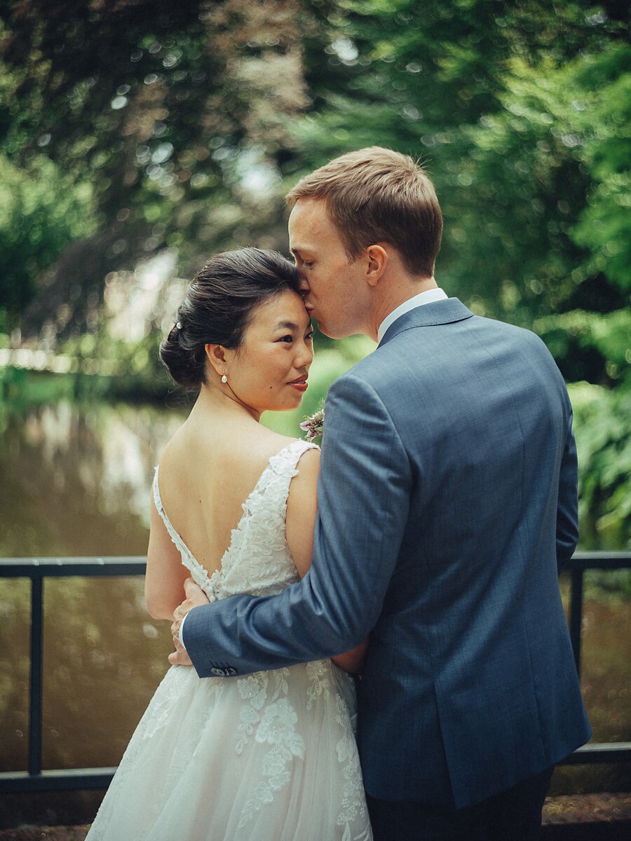 Wedding SP26 Bruidegom in blauw pak kust het voorhoofd van zijn bruid bij een groene grachtenleuning, haar kanten rug in het licht, een teder portret