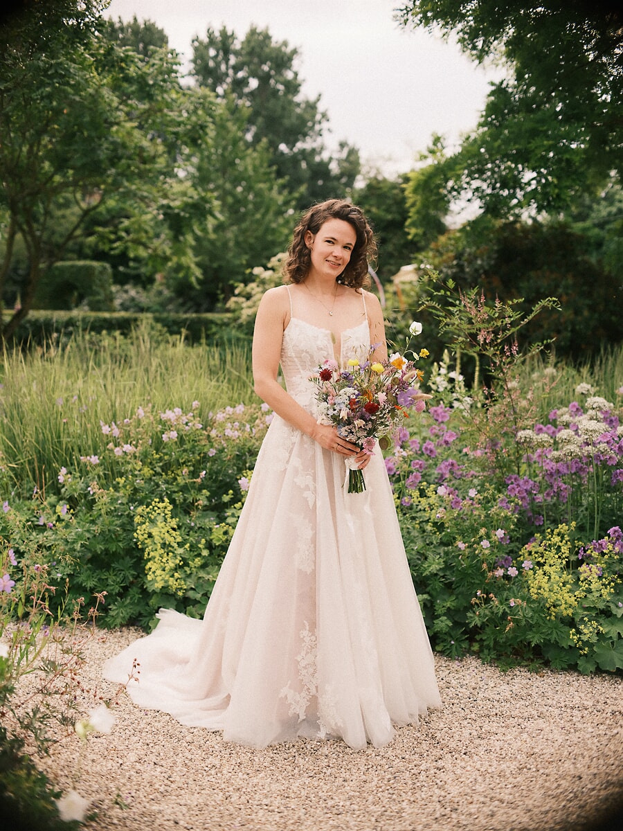 Wedding SP3 Met een veldboeket op een grindpad in een zomerse tuin lacht de bruid naar de trouwfotograaf in Amsterdam