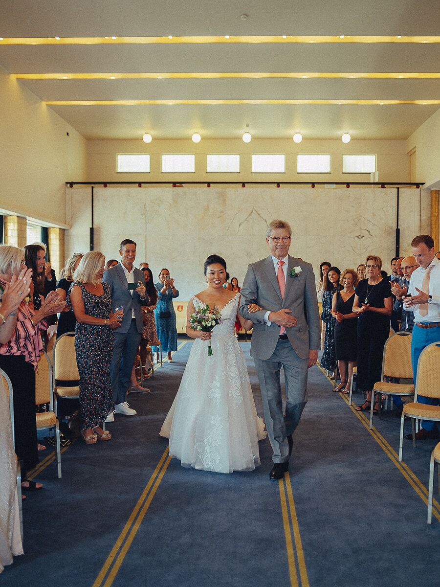 Wedding SP31 Arm in arm met haar vader loopt de bruid door het blauwe gangpad terwijl gasten van beide kanten applaudisseren, een documentaire ceremoniefoto