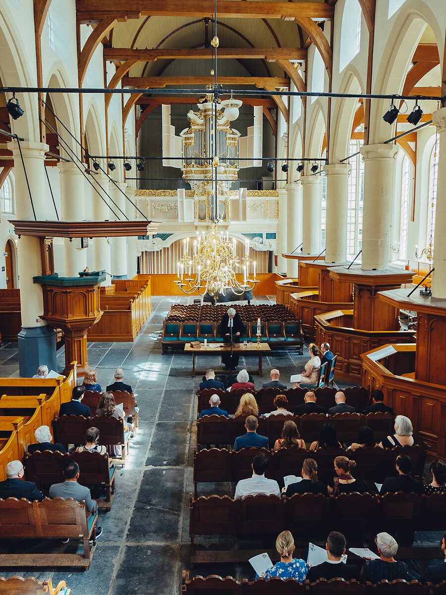Wedding SP32 Historische Nederlandse kerk met houten banken en vergulde kroonluchter loopt vol gasten, een groots beeld van een trouwfotograaf in Amsterdam