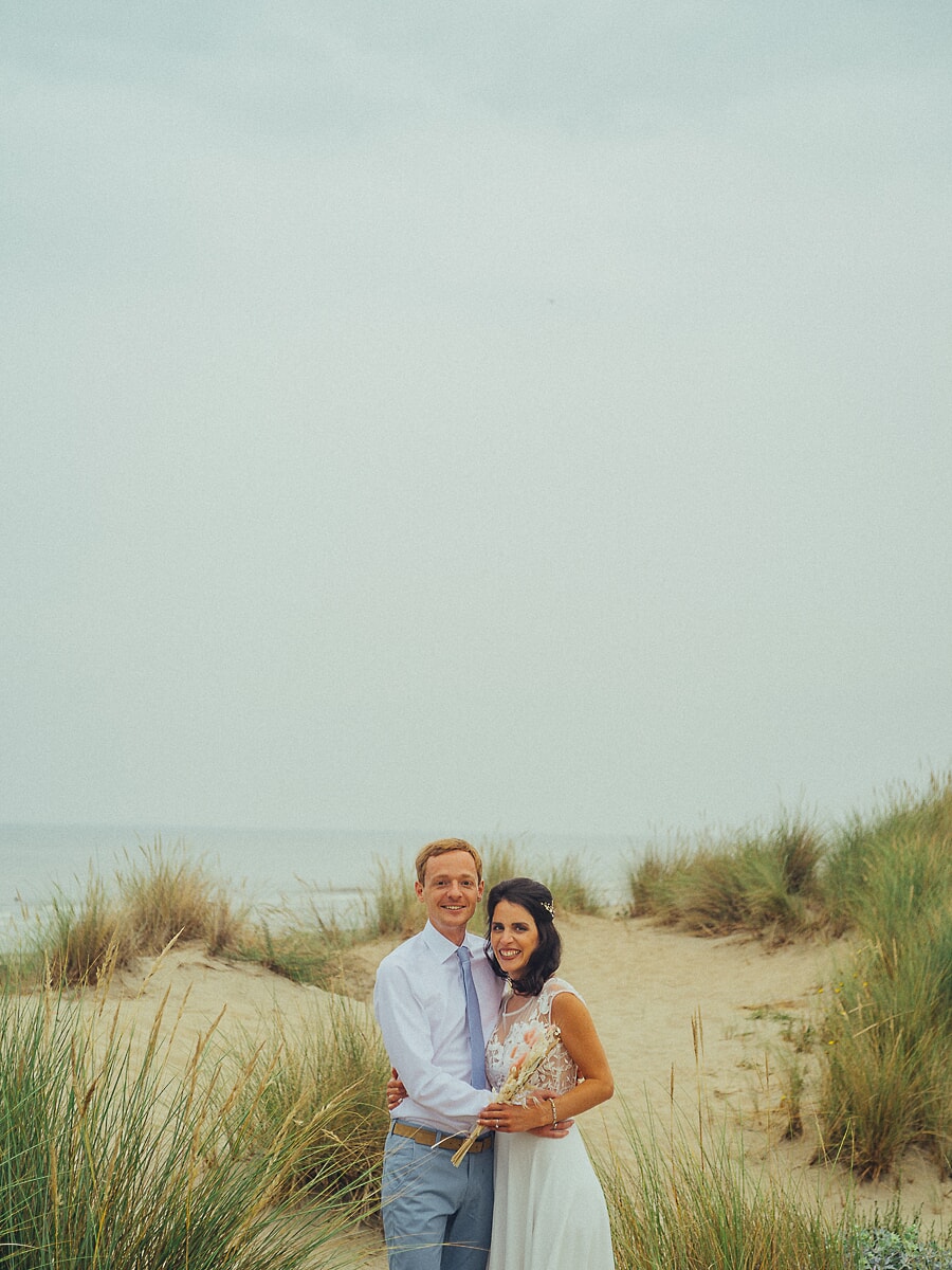 Wedding SP33 Stel omhelst elkaar tussen het helmgras in zacht kustlicht, de bruid met een droogbloemenboeket, een ontspannen strandportret