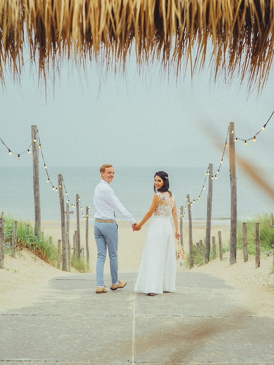 Wedding SP34 Onder lichtslingers kijken de pasgetrouwden om terwijl ze naar de duinen lopen, gefotografeerd door een trouwfotograaf in Amsterdam