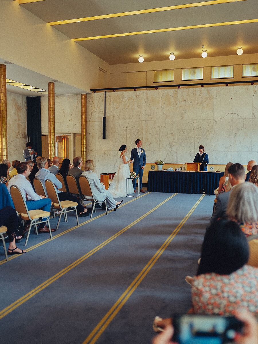 Wedding SP35 Bruid en bruidegom staan voor de ambtenaar in een marmeren zaal, gasten kijken toe vanaf gouden stoelen, een documentaire huwelijksfoto