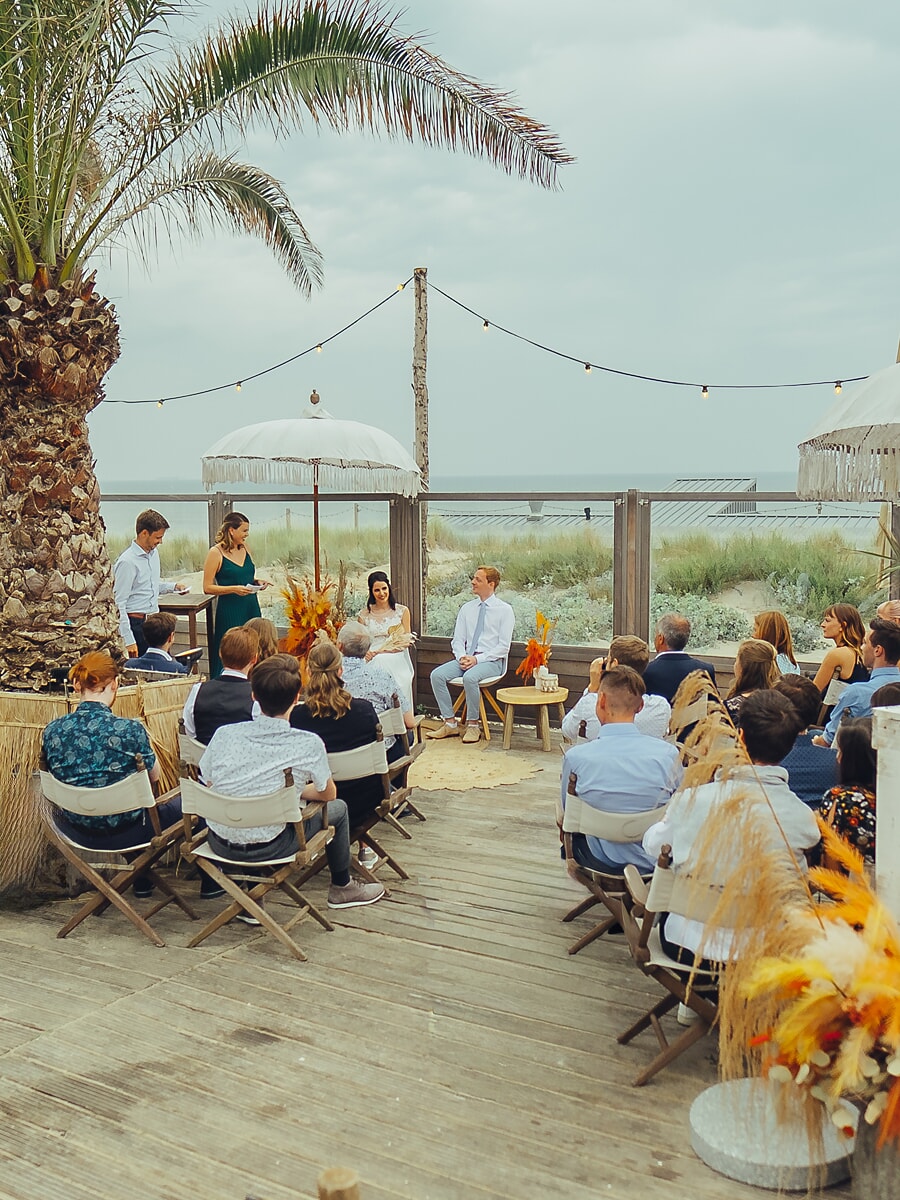 Wedding SP36 Gasten op een houten stranddek kijken naar het zittende stel onder een parasol met franjes, vastgelegd door een trouwfotograaf in Amsterdam