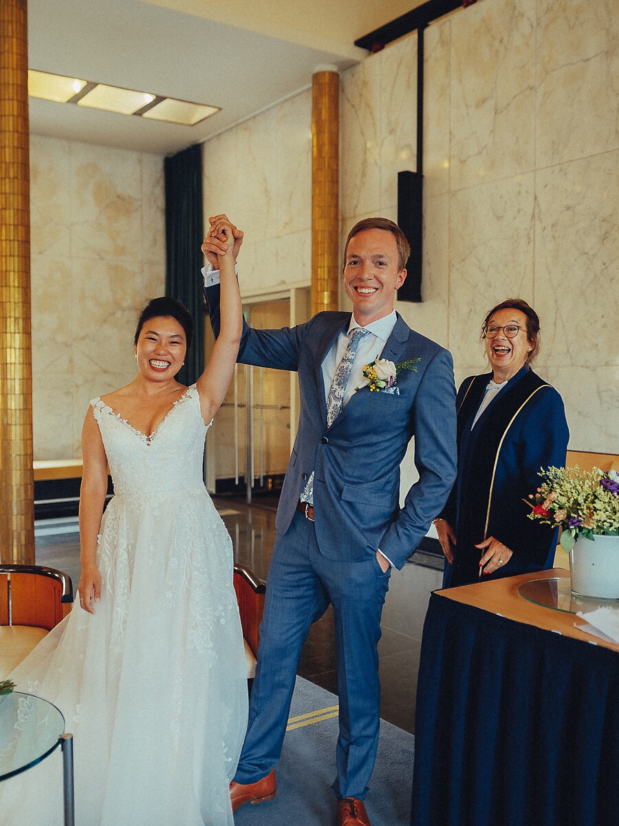 Wedding SP37 Lachende bruidegom heft de hand van zijn bruid triomfantelijk omhoog naast de ambtenaar in een marmeren stadhuis, een blije ceremoniefoto