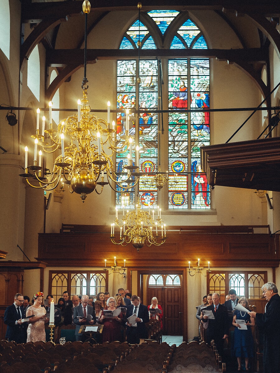 Wedding SP38 Onder glas-in-lood en koperen kronen staan gasten met liedboekjes in de hand, een tijdloos beeld van een trouwfotograaf in Amsterdam