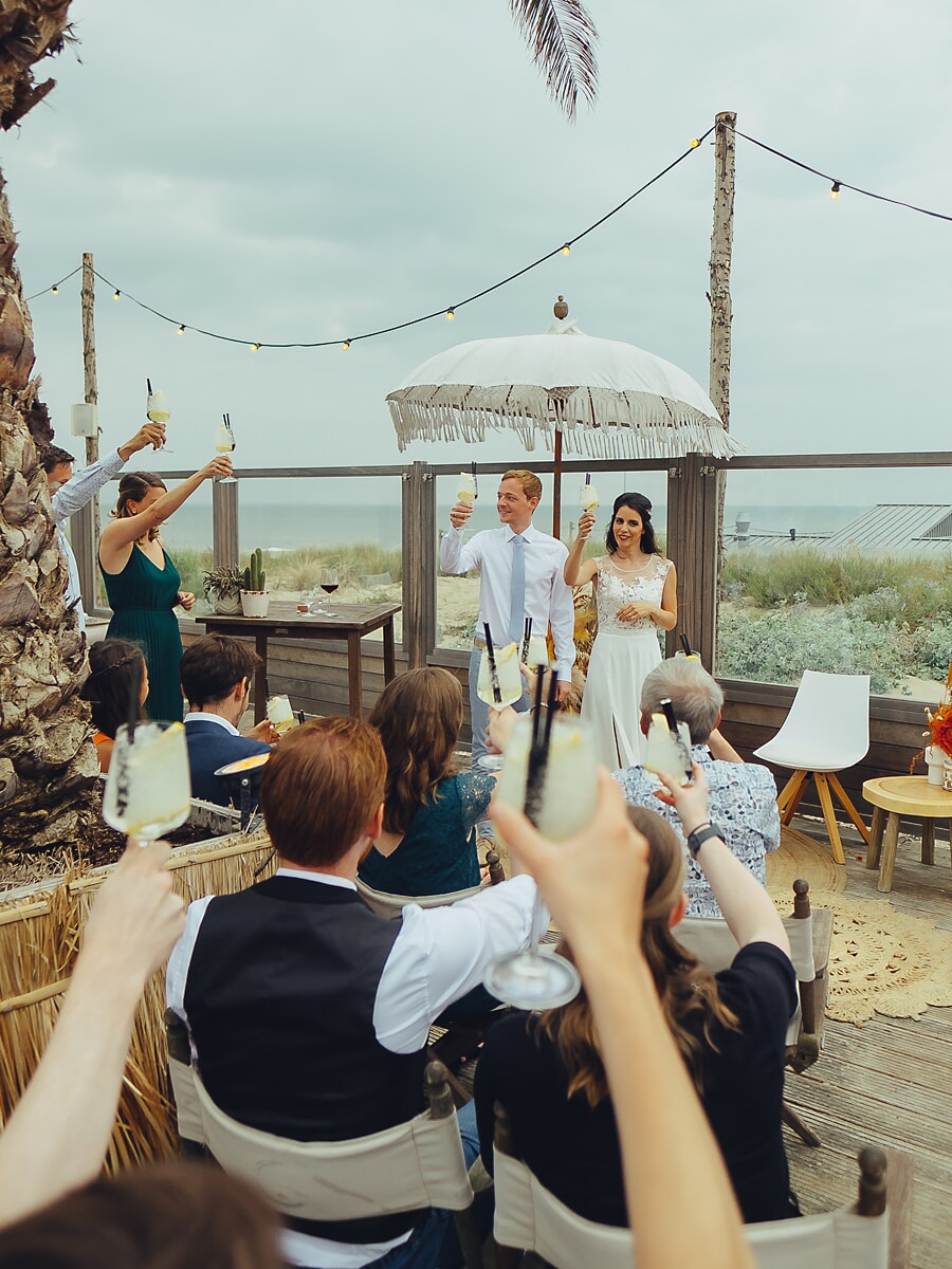 Wedding SP39 Met geheven glazen richting het stel onder een parasol bij een strandlocatie proosten de gasten, een levendige bruiloftsfoto