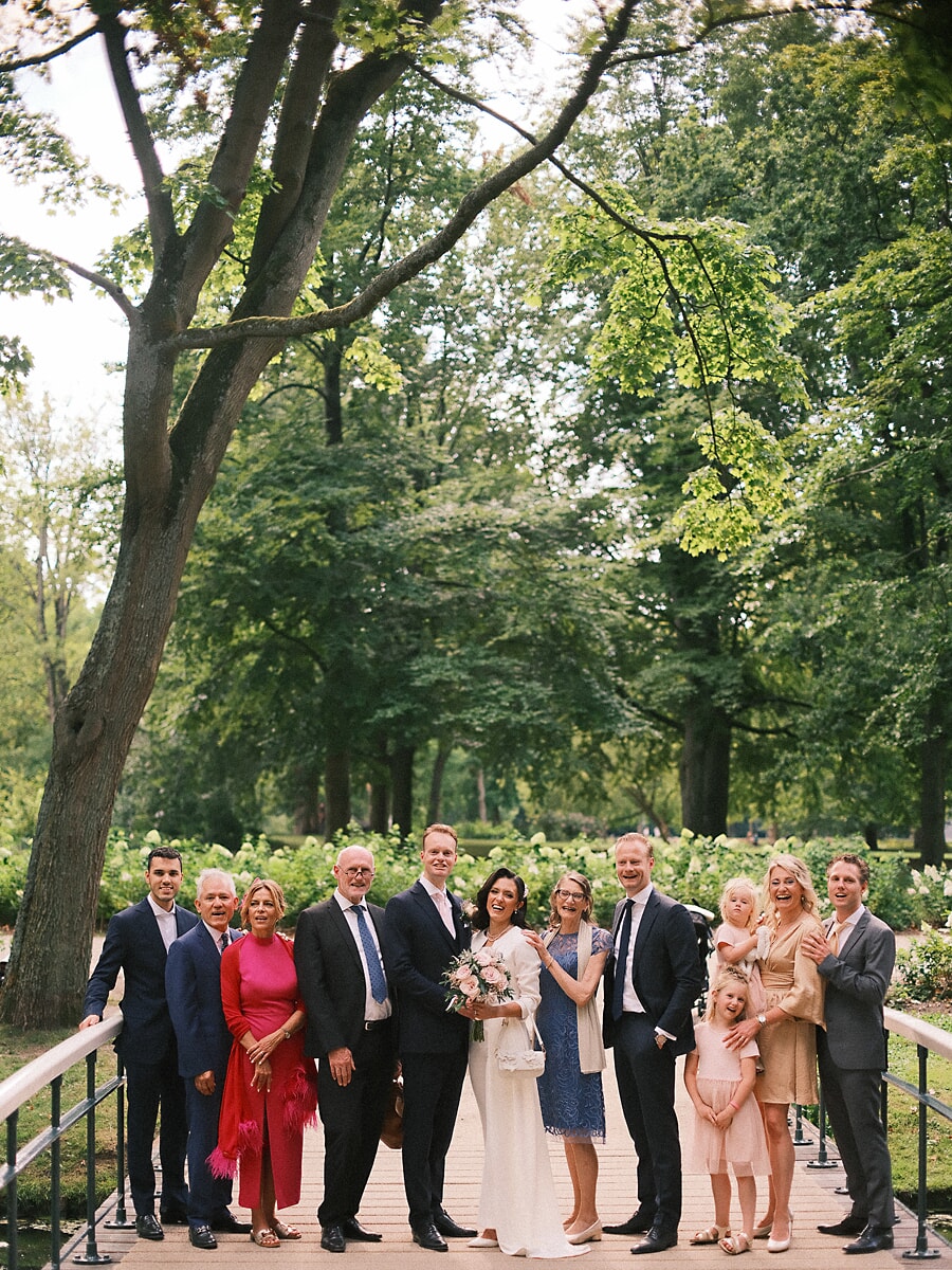 Wedding SP4 Familie en vrienden verzamelen zich rond het bruidspaar op een houten parkbrug, een vrolijk groepsportret uit de trouwreportage