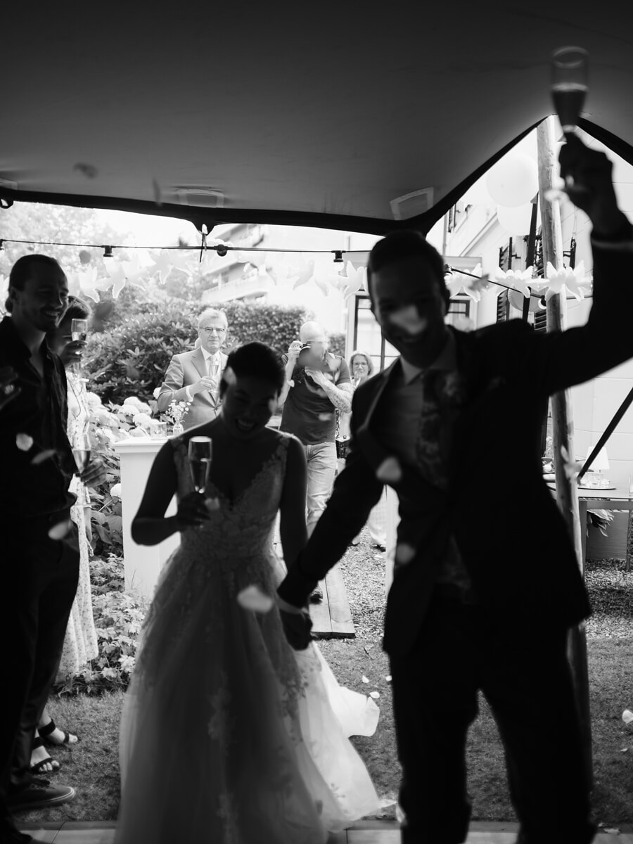 Wedding SP40 Zwart-witfoto van de pasgetrouwden die met champagne naar buiten stappen terwijl gasten juichen, van een trouwfotograaf in Amsterdam