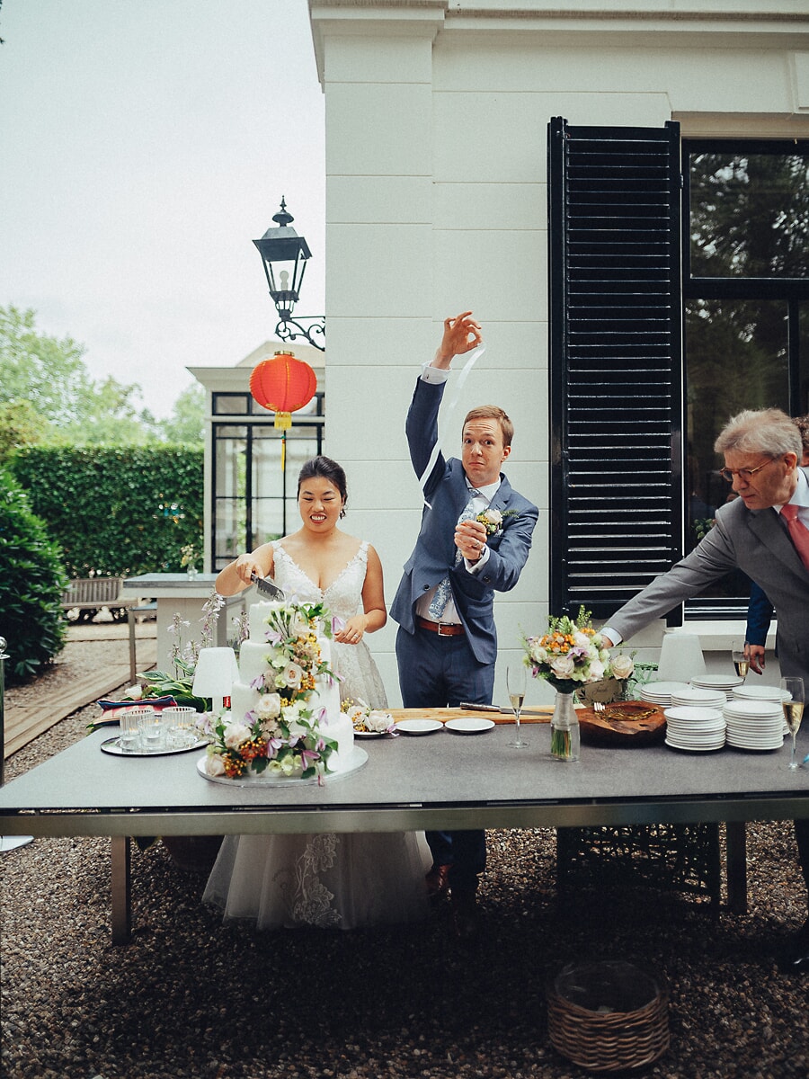 Wedding SP41 Bruidegom heft het mes boven zijn hoofd terwijl zijn bruid de bloementaart aansnijdt op het terras, een speelse feestfoto
