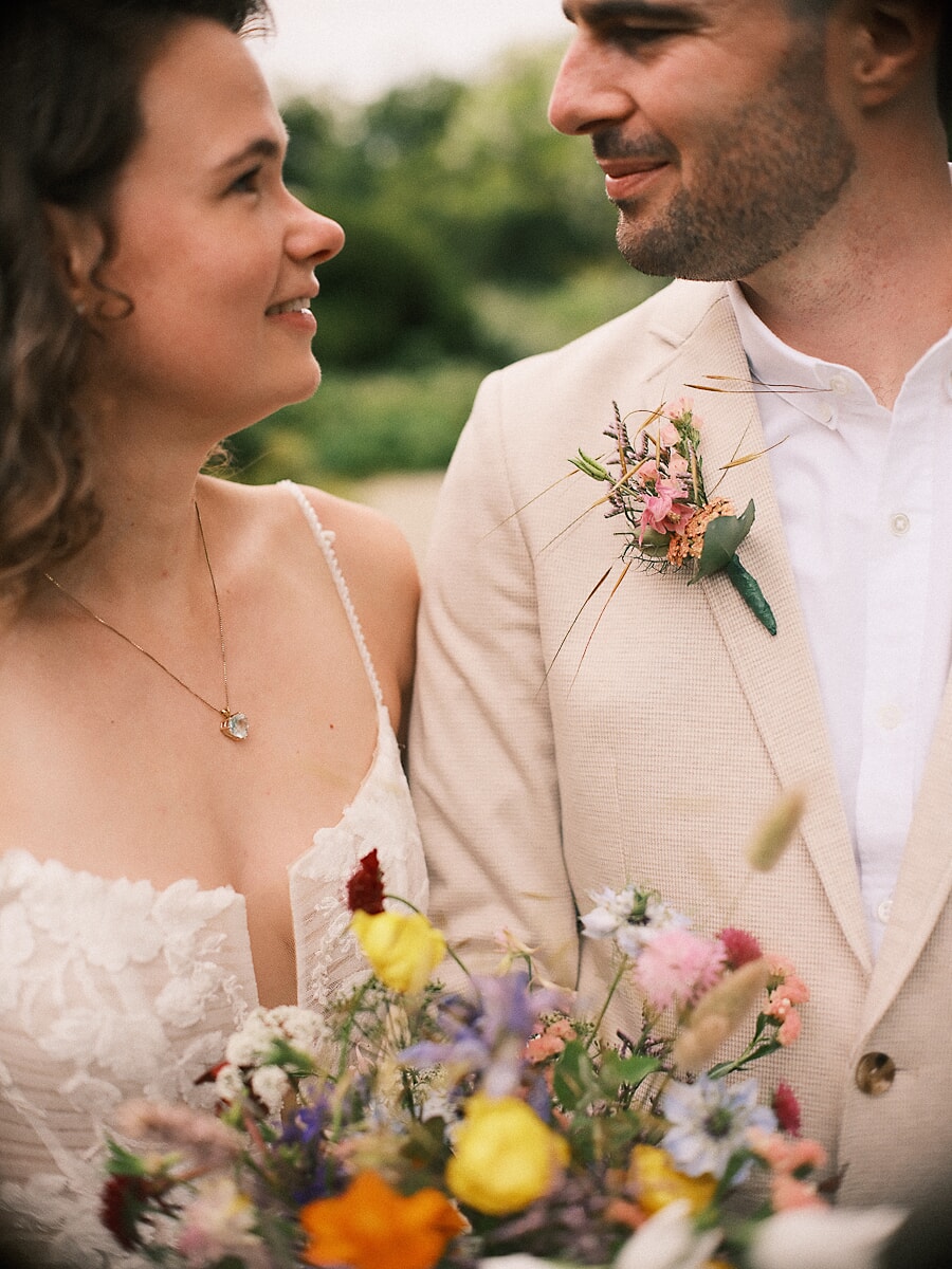 Wedding SP5 Bruid en bruidegom kijken elkaar van opzij aan, droogbloemen op zijn linnen revers en gele bloemen in haar boeket, een intieme bruiloftsfoto