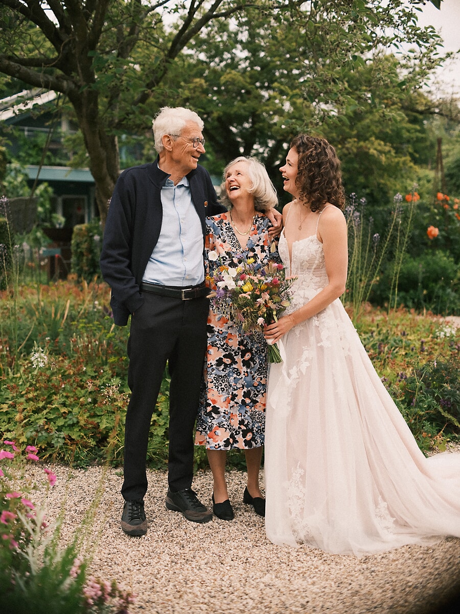 Wedding SP7 Arm in arm met haar ouders houdt de lachende bruid haar veldboeket vast, een hartverwarmend beeld van een trouwfotograaf in Amsterdam