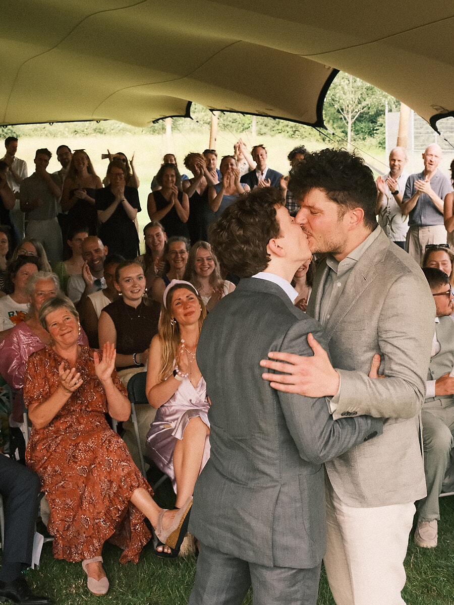 Wedding SP8 Twee bruidegoms kussen elkaar onder een tent terwijl gasten applaudisseren, een spontane foto van een gelijkgeslachtelijk huwelijk