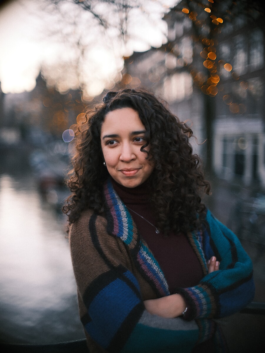 Vrouw met donkere krullen en over elkaar geslagen armen in een gestreept vest, met lichtjes achter haar in de schemering