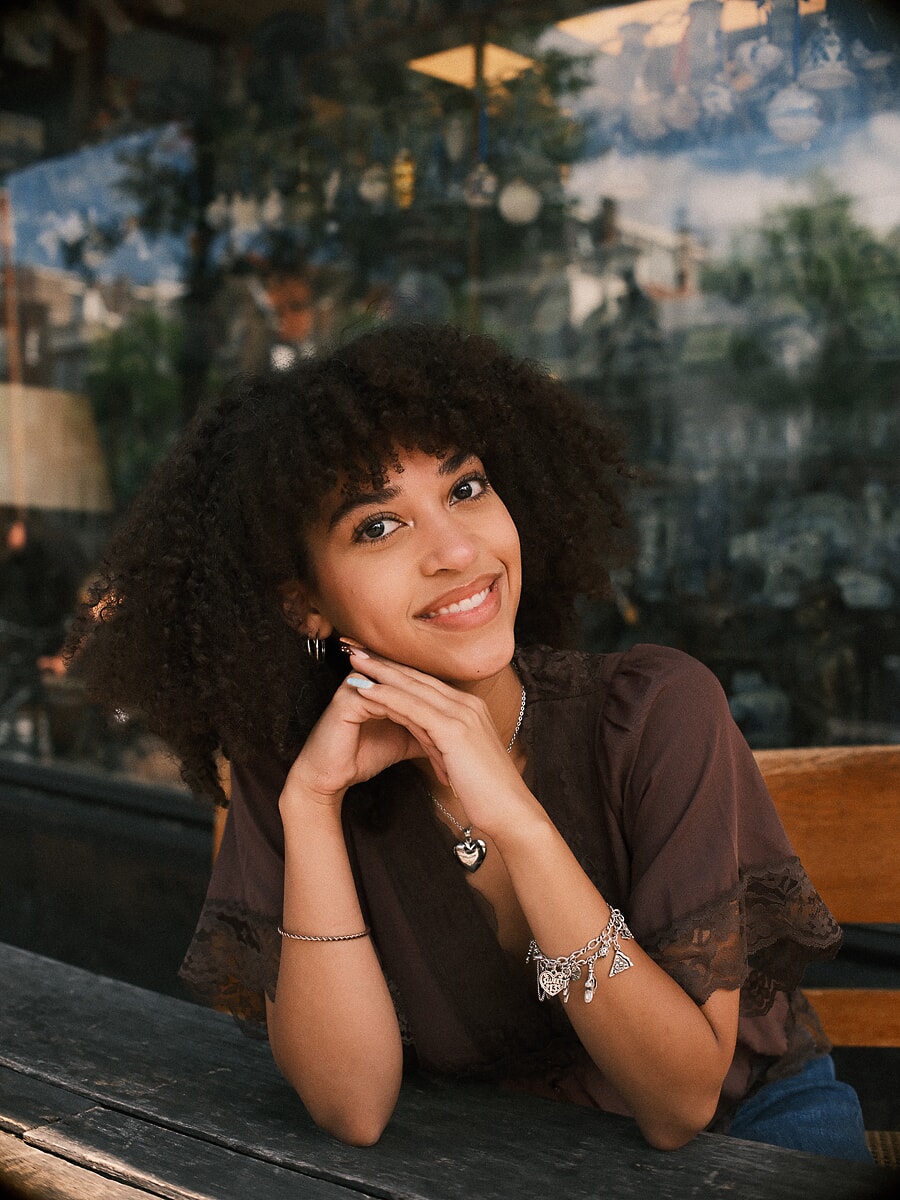 Vrouw met haar kin op haar handen aan een cafétafel voor een geschilderd grachtentafereel, een lifestyleportret in een Amsterdams café