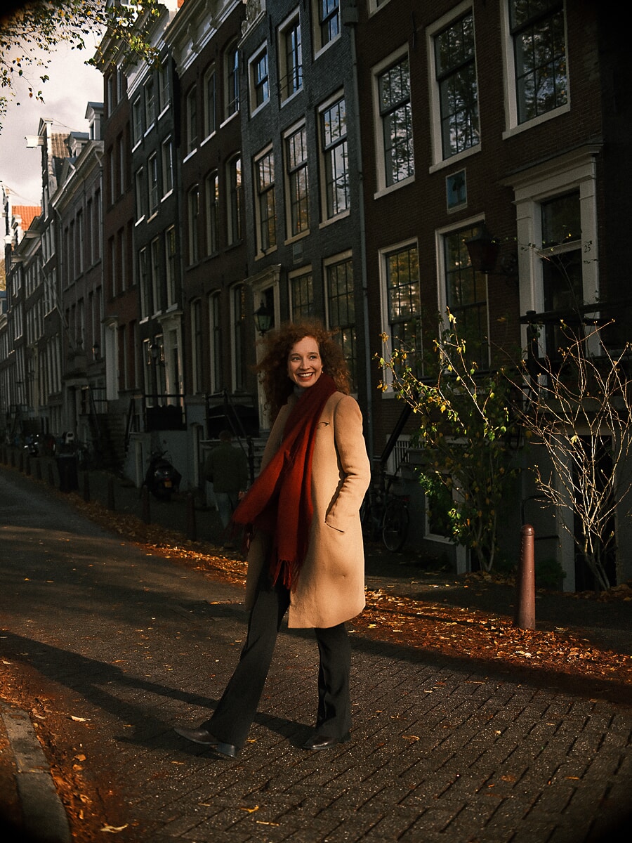 Vrouw in kameelkleurige jas en rode sjaal in een herfstige straat langs een gracht vol bladeren, een herfstportret in Amsterdam