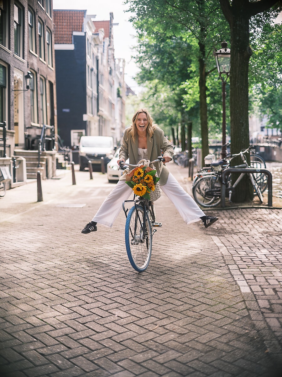 Vrouw rolt met gestrekte benen lachend op de camera af, met zonnebloemen aan het stuur, een straatportret in Amsterdam
